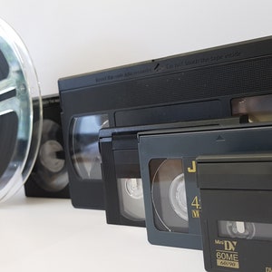 Può includere: Una collezione di cassette VHS vintage e un rotolo di pellicola. Le cassette sono nere e hanno etichette con il testo "Non mettere il nastro in modalità sicura" e "Mini DV 60ME 640x480". Il rotolo di pellicola è in plastica trasparente con una pellicola nera all'interno.