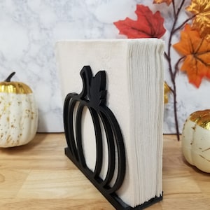 May include: Black pumpkin-shaped napkin holder made of wood. The holder is filled with white napkins.