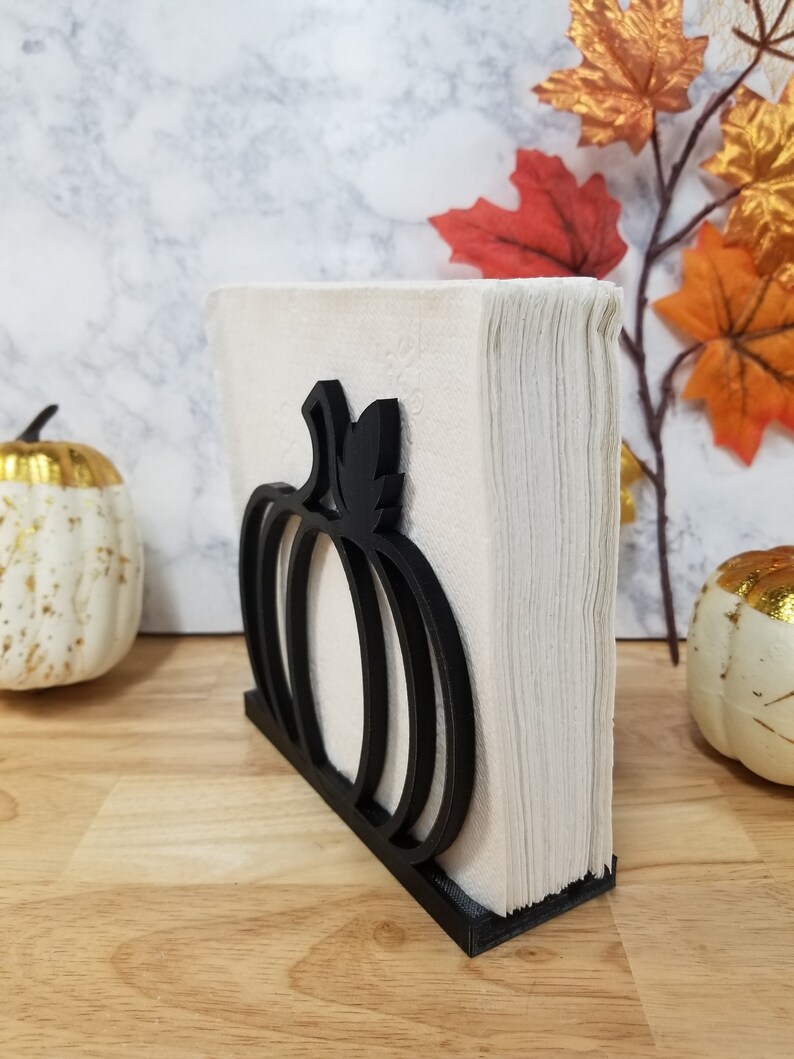 May include: Black pumpkin-shaped napkin holder made of wood. The holder is filled with white napkins.