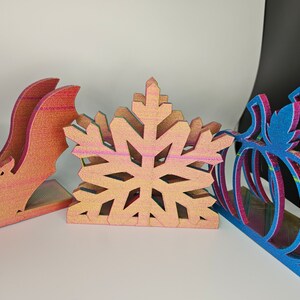 May include: Three wooden napkin holders, one shaped like a bat, one like a snowflake, and one like a pumpkin. The holders are all painted in a rainbow iridescent finish.