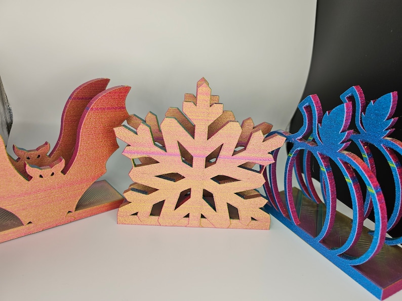 May include: Three wooden napkin holders, one shaped like a bat, one like a snowflake, and one like a pumpkin. The holders are all painted in a rainbow iridescent finish.