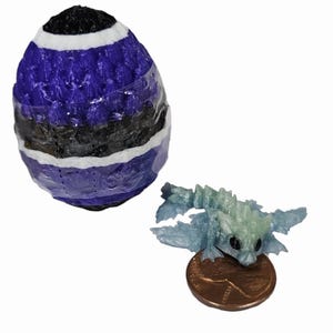 May include: A purple and black striped egg with a small, light blue dragon figurine next to it. The dragon is sitting on a penny for scale.