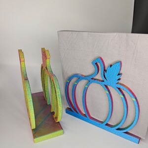 May include: Two iridescent pumpkin-shaped napkin holders. One is blue with a green leaf and the other is yellow, orange, and green.