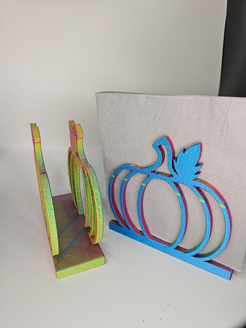 May include: Two iridescent pumpkin-shaped napkin holders. One is blue with a green leaf and the other is yellow, orange, and green.