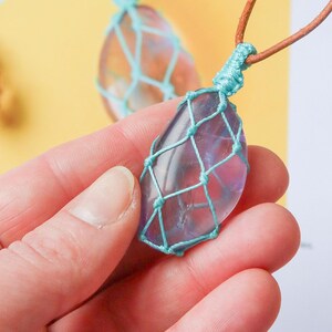 May include: A purple crystal pendant wrapped in a turquoise macrame net, hanging from a brown leather cord. The pendant is being held in a hand, with a printed card in the background that says "Net Guide Macrame Pendant Pattern".