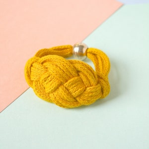 Pu&ograve; includere: Un braccialetto intrecciato giallo con un accento di perline d'argento. Il braccialetto &egrave; su uno sfondo blu chiaro e rosa.