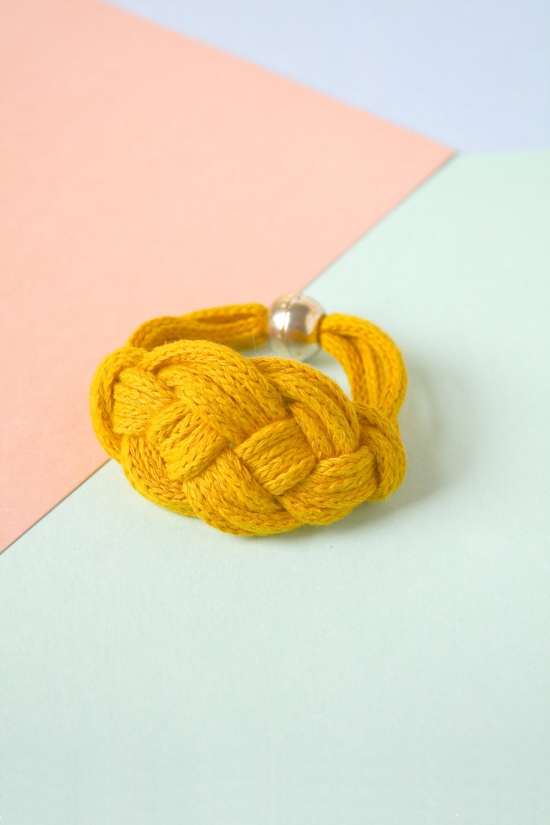 Pu&ograve; includere: Un braccialetto intrecciato giallo con un accento di perline d'argento. Il braccialetto &egrave; su uno sfondo blu chiaro e rosa.