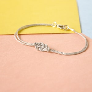 Pu&ograve; includere: Un braccialetto d'argento con un design annodato su uno sfondo rosa chiaro e giallo.