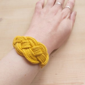 Pu&ograve; includere: Un braccialetto intrecciato giallo fatto di corda spessa. Il braccialetto &egrave; indossato al polso.