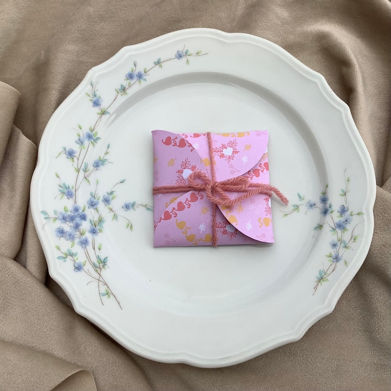 May include: A pink and white patterned gift box with a pink ribbon tied around it. The box is sitting on a white plate with a floral design.