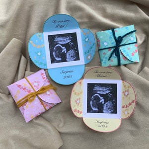 May include: Two yellow, blue, and pink cardstock envelopes with ultrasound images and the text "Tu vas &ecirc;tre Papy!" and "Tu vas &ecirc;tre Mamie!"  The envelopes are tied with twine and ribbon.