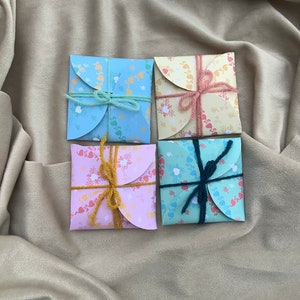 May include: Four small, folded, paper envelopes with a heart and floral pattern. Each envelope is tied with a different colored ribbon. The envelopes are on a light brown fabric background.
