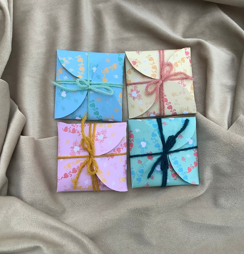 May include: Four small, folded, paper envelopes with a heart and floral pattern. Each envelope is tied with a different colored ribbon. The envelopes are on a light brown fabric background.