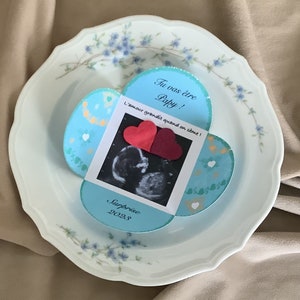 May include: A blue and white card with a heart-shaped opening reveals a sonogram image and two red hearts. The card reads "Tu vas &ecirc;tre Papy! L'amour grandit quand on aime! Surprise 2023."