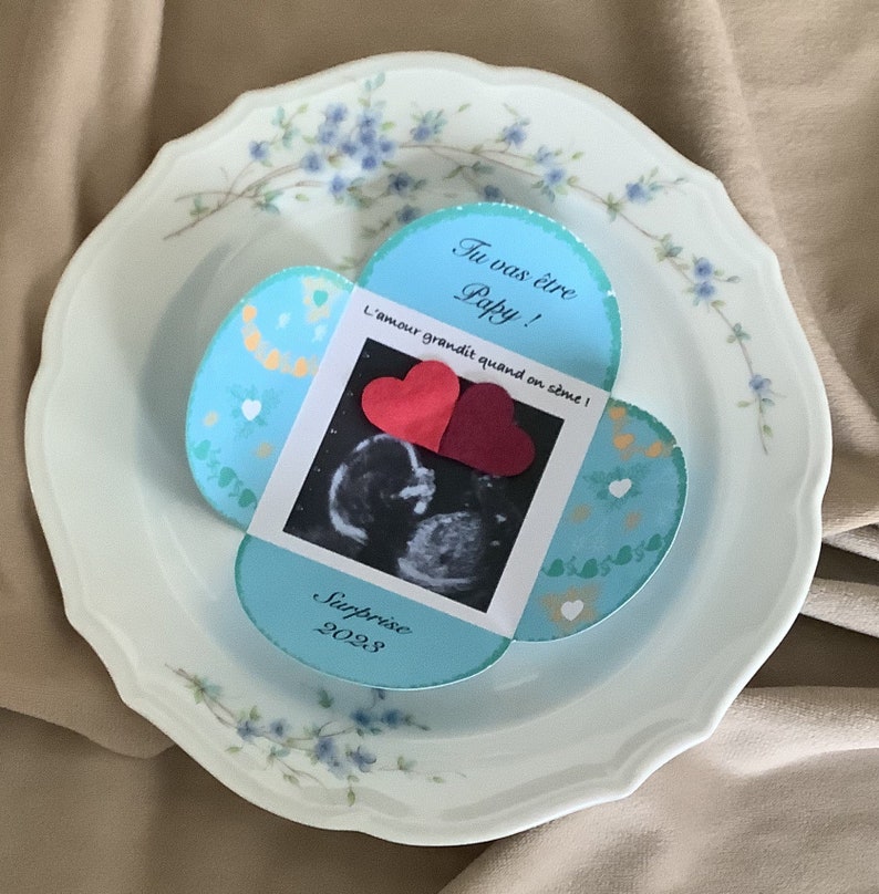 May include: A blue and white card with a heart-shaped opening reveals a sonogram image and two red hearts. The card reads "Tu vas &ecirc;tre Papy! L'amour grandit quand on aime! Surprise 2023."