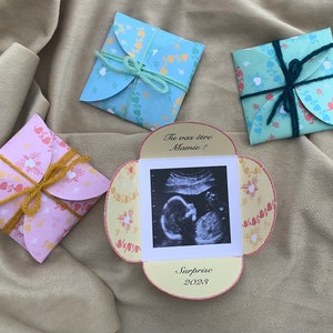 May include: A yellow, pink, and blue card with a heart pattern. The card has a small ultrasound image and the text "Tu vas &ecirc;tre Mamie! Surprise 2023".