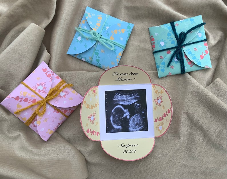May include: A yellow, pink, and blue card with a heart pattern. The card has a small ultrasound image and the text "Tu vas &ecirc;tre Mamie! Surprise 2023".