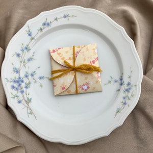 May include: A white porcelain plate with a floral design featuring blue flowers and green stems. A small, folded, yellow and red patterned paper gift box with a brown twine bow sits in the center of the plate.