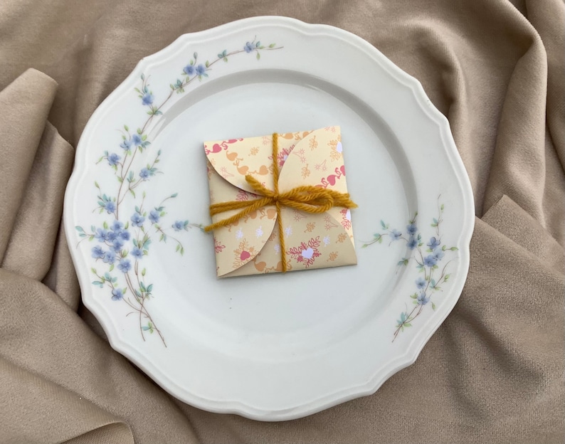 May include: A white porcelain plate with a floral design featuring blue flowers and green stems. A small, folded, yellow and red patterned paper gift box with a brown twine bow sits in the center of the plate.