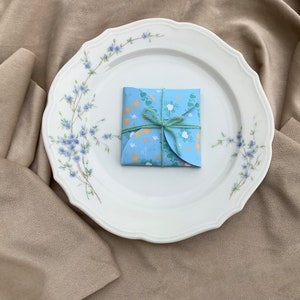 May include: A blue and white patterned gift box tied with a green ribbon, sitting on a white plate with a floral design. The plate is on a beige fabric surface.