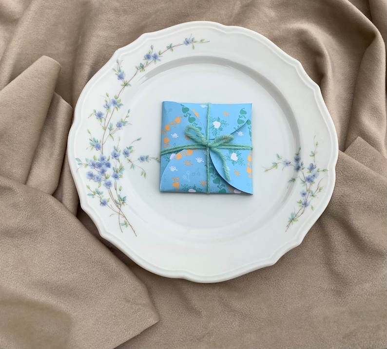 May include: A blue and white patterned gift box tied with a green ribbon, sitting on a white plate with a floral design. The plate is on a beige fabric surface.