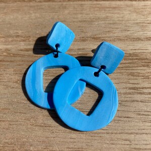 May include: A pair of blue earrings with a square shape and a circle shape with a square cutout. The earrings are made of polymer clay and have a marbled effect.