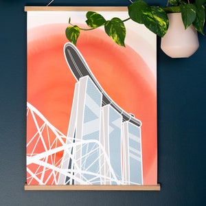 May include: A print of a modern architectural design, featuring a bridge and tall buildings against a red and orange background. The print is held by a wooden frame and suspended by a white string. Green plants in white pots hang on either side.