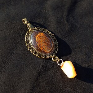 Peut inclure: Un pendentif doré avec un motif de peau de serpent brun et orange à l'intérieur d'un ovale transparent. Le pendentif est attaché à une chaîne dorée avec une petite perle orange clair.