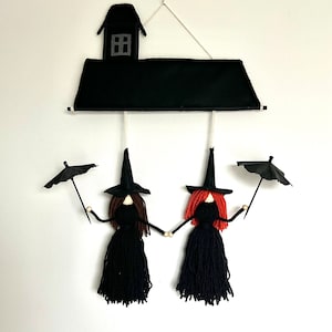 Puede incluir: Una decoración de Halloween caprichosa con dos muñecas de hilo vestidas de brujas, tomadas de la mano. Llevan vestidos y sombreros negros, una tiene el pelo castaño y la otra rojo. Una casa negra y paraguas completan la escena.