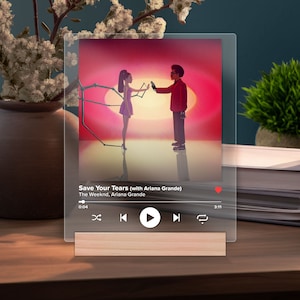 May include: A digital music player screen displaying the song "Save Your Tears (with Ariana Grande)" by The Weeknd and Ariana Grande. The album art features a man and woman standing in front of a red and orange sunset.