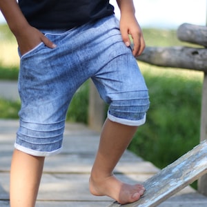 Może przedstawiać: Dziecko w niebieskich dżinsowych spodenkach z białym wykończeniem. Spodenki mają przetarty wygląd i są podwinięte na dole.