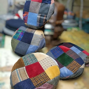 Può includere: Quattro cappelli newsboy con design patchwork. Ogni cappello presenta una combinazione unica di tessuti in vari colori, tra cui velluto a coste, tweed e scozzese. I cappelli sono disposti in modo leggermente sovrapposto, mostrando i loro design individuali.