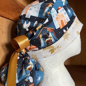 May include: A scrub cap featuring a dark blue fabric with a repeating nativity scene print, including figures in a stable and architectural structures. The cap has a white band with gold snowflake patterns and a solid gold ribbon tied at the back.