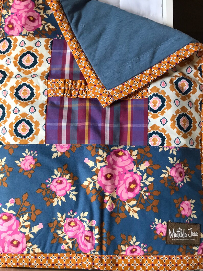 May include: A blue and brown patchwork quilt with a floral pattern and a Matilda Jane label. The quilt has a blue backing and a brown and white patterned border.