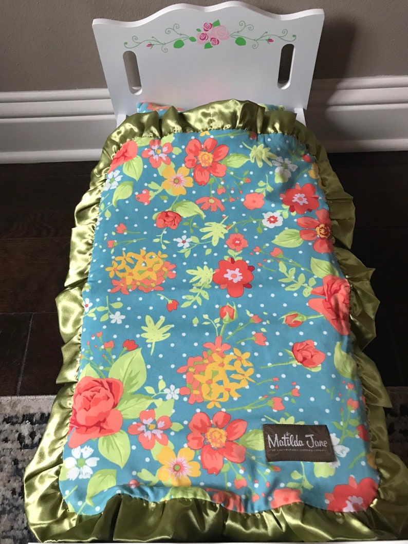 May include: A white doll bed with a blue and white floral patterned blanket with green trim and a Matilda Jane label. The blanket has a satin border.