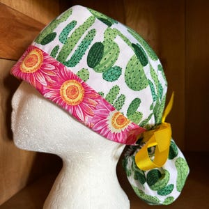 May include: A white scrub cap with a pattern of green cacti. The cap has a pink floral band with yellow centres and a yellow ribbon tie. The cap is displayed on a white mannequin head.