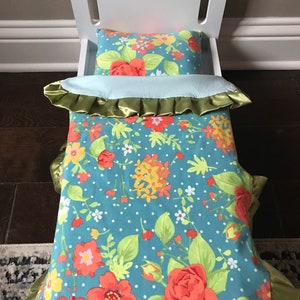 May include: A white doll bed with a floral patterned comforter and pillow. The comforter has a green satin trim and the pillow has a blue background with red, yellow, and white flowers.