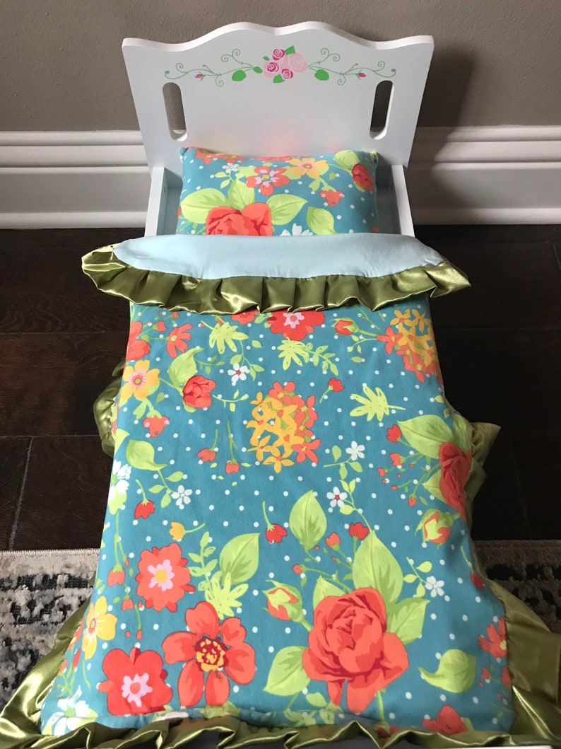 May include: A white doll bed with a floral patterned comforter and pillow. The comforter has a green satin trim and the pillow has a blue background with red, yellow, and white flowers.