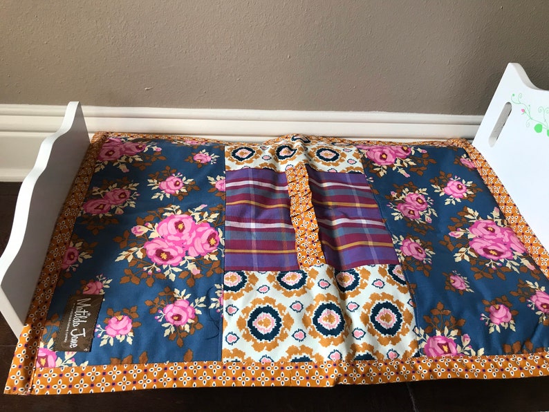 May include: A blue and white patchwork quilt with pink flowers and a brown border. The quilt has a "Matilda Jane" label.