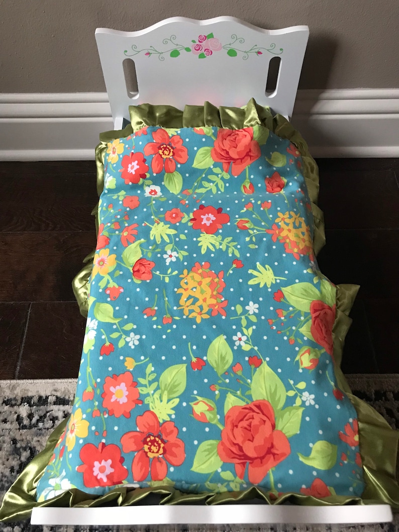 May include: A white doll bed with a floral patterned bedding set. The bedding has a blue background with red, yellow, and green flowers. The bedding has a green ruffled trim.