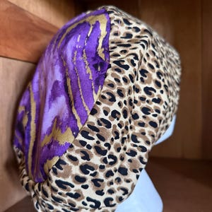 May include: A patterned scrub cap featuring a combination of purple and gold abstract design and a leopard print pattern. The cap is designed to fit comfortably and securely, offering a stylish and functional accessory for medical professionals.