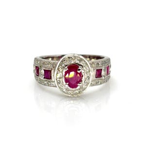 Peut inclure: Bague en argent avec une grosse pierre centrale ovale en rubis, entourée d'un halo de petits diamants. La bande comporte des rubis carrés et de petits diamants. La bague est posée sur fond blanc.