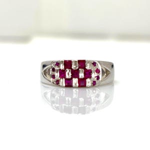 May include: A silver ring featuring a rectangular arrangement of square, red gemstones. The ring has a polished finish and a geometric pattern of alternating red stones. The ring is displayed on a white surface.