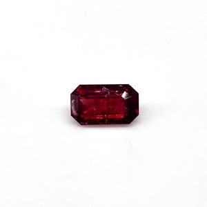 Puede incluir: Una gema roja oscura de corte rectangular con facetas. La gema se encuentra sobre un fondo blanco, resaltando su rico color y claridad. La forma de la gema es un corte esmeralda, con una superficie ligeramente imperfecta.