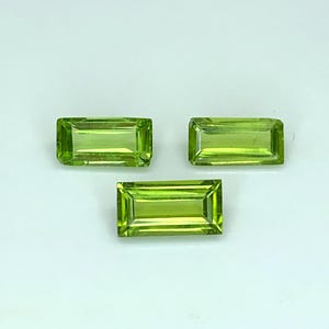 May include: Three loose, rectangular, baguette-cut peridot gemstones. Each stone features a vibrant, translucent lime-green color with faceted surfaces that reflect light, arranged in a triangular formation against a plain, light background.