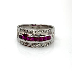 Peut inclure: Une bague en métal argenté dotée d'une rangée centrale horizontale de pierres carrées d'un rouge rosé vif. La rangée centrale est bordée de deux bandes plus fines incrustées de petites pierres claires et scintillantes, créant un aspect multicouche.