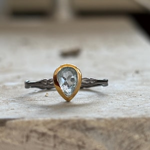 May include: A close-up of a ring featuring a pear-shaped, light blue gemstone set in a gold bezel. The band is dark gray with a decorative pattern. The ring is set on a light-colored surface.
