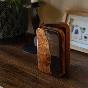 Może przedstawiać: Drewniane pudełko z otwieraną pokrywą, z połączeniem jasnych i ciemnych odcieni drewna. Pudełko ma błyszczące wykończenie i mały, ciemny okrągły detal. Tło obejmuje oprawiony obraz i dekoracyjną roślinę.