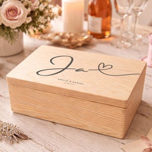 Puede incluir: Una caja de madera de color claro con un diseño decorativo en la tapa. La caja está personalizada con un nombre y un símbolo de corazón. La escena incluye flores, champán y otros accesorios temáticos de boda.