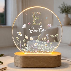 Pode incluir: Uma luz noturna circular de acrílico com uma borda de brilho quente, apresentando um design floral, uma letra E grande e o nome Emilia impresso em branco. A luz está montada numa base de madeira clara, lisa e oval, com um cabo de alimentação.
